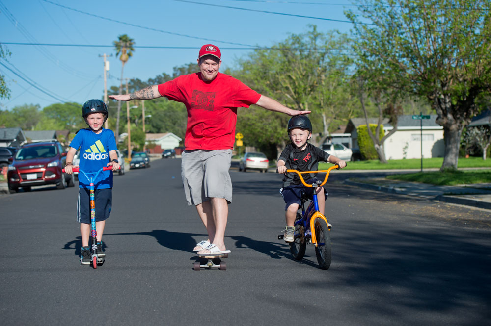 Father riding skateboard next to son on bicycle
